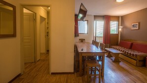 A room with a wooden dining table, chairs, a sofa, and a TV mounted on the wall. - Près du centre & des pistes | Charmant T3 de 55m² (Orcières)