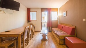 A compact room with a wooden dining table, chairs, a sofa, and a television mounted on the wall. - Près du centre & des pistes | Charmant T3 de 55m² (Orcières)