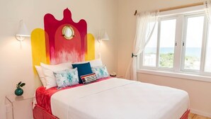 Un lit avec une tête de lit rouge, une literie blanche et une vue sur la mer par la fenêtre.
