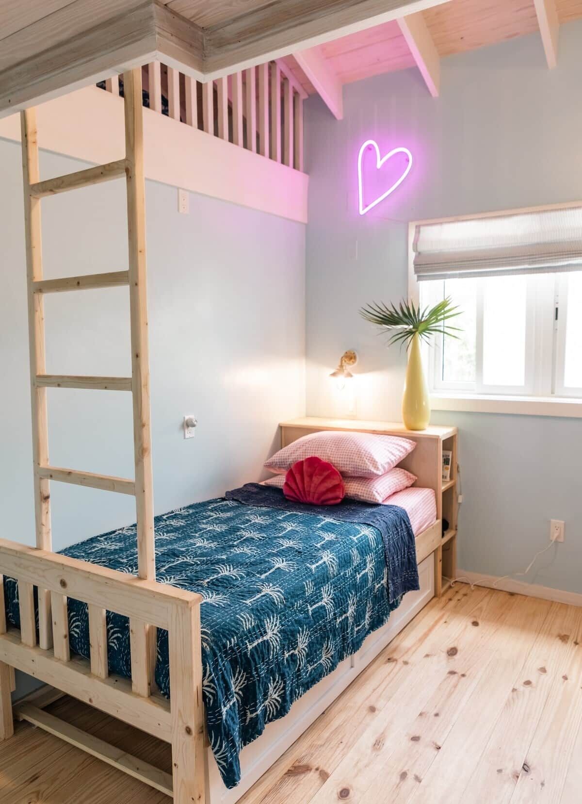 A bunk bed with a ladder, a bedside table with a lamp, a vase with a plant, and a window with a view.