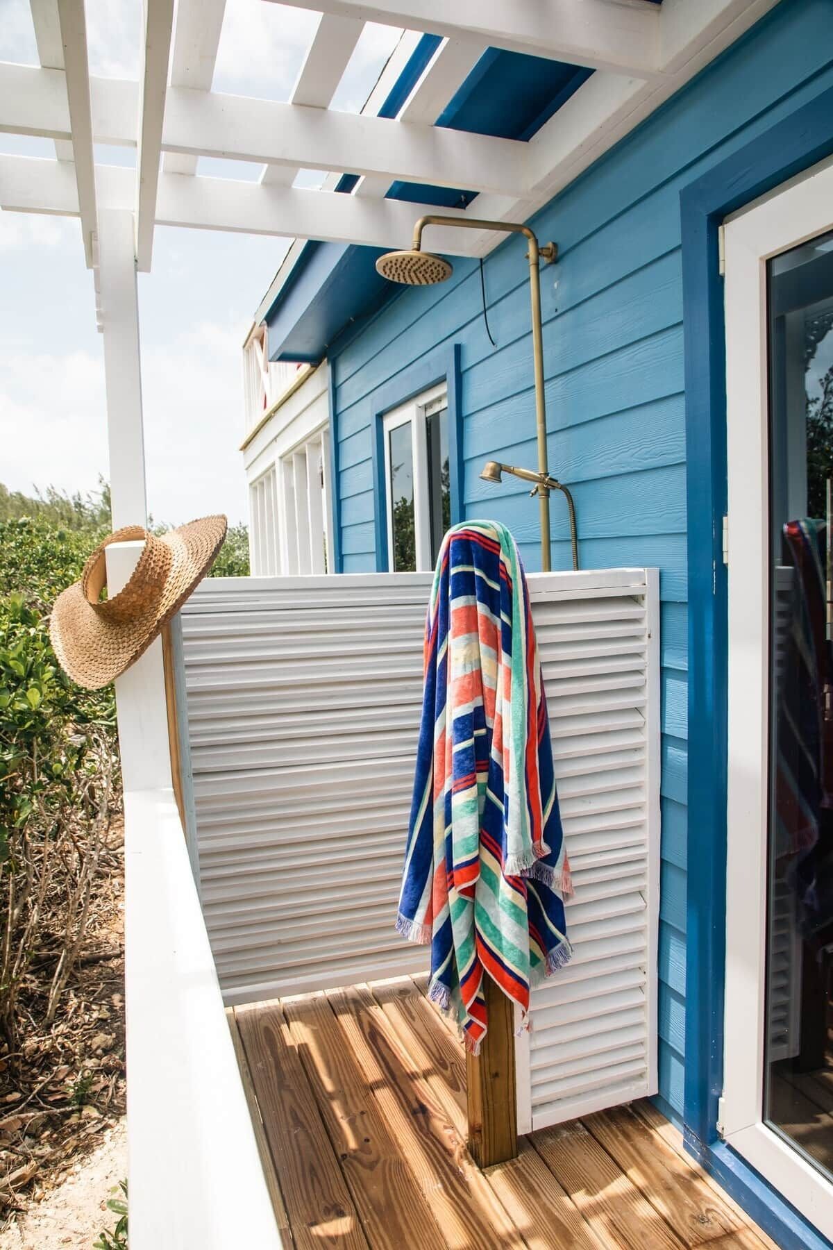 Une maison bleue avec un porche blanc, une terrasse en bois, un chapeau de paille et une serviette à rayures.