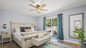A bedroom with a wooden bed, a ceiling fan, a nightstand, a bench, a large mirror, and a view of the outdoors. - Tahoe Getaway | 2 King Suites & Hot Tub | Near Town, Lake Activities & Trails (Truckee)