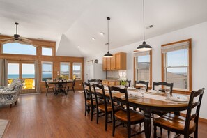 A spacious dining area with a large wooden table set for six, surrounded by wooden chairs. There are large windows offering a view of the ocean.