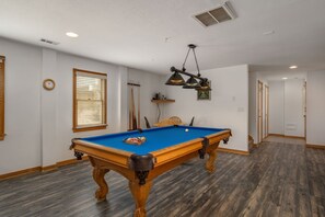 A pool table with a blue felt surface, wooden legs, and a corner pocket. There is a clock on the wall and a window with blinds.