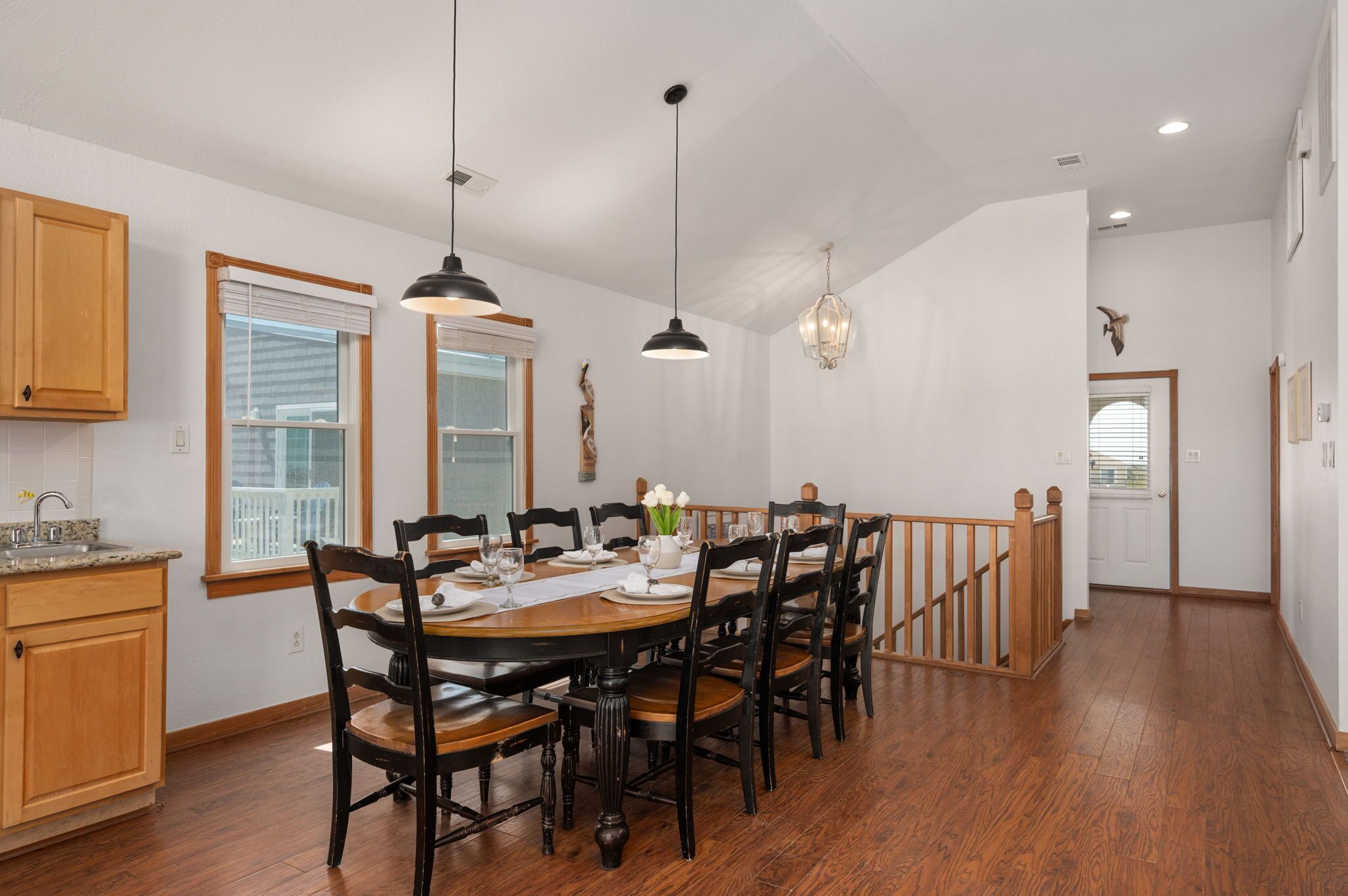 Uma sala de jantar com uma mesa de madeira posta para seis pessoas, cercada por cadeiras. Há luminárias pendentes sobre a mesa e um candelabro ao fundo. Uma escada de madeira com corrimão leva a outro cômodo.