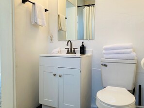 Un baño con un lavamanos blanco, grifo negro y un inodoro. Hay toallas colgadas en un perchero y un espejo sobre el lavamanos.