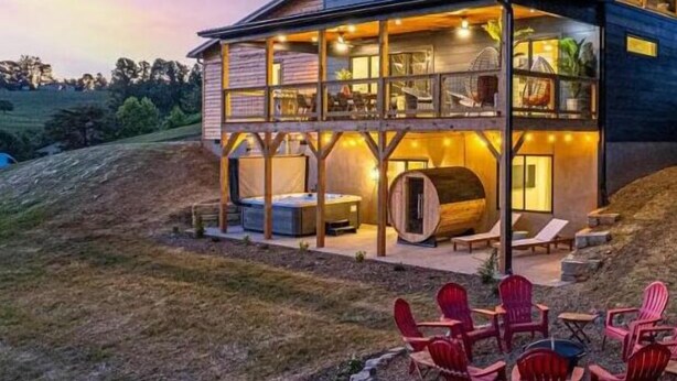 A two-story wooden house with a balcony, outdoor seating, and a view of the surrounding landscape. - Symphony of Luxury|Hot Tub+Game Room+Sauna+Views (Leicester)