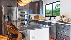 A modern kitchen with dark wood cabinets, stainless steel appliances, a large island with bar stools, and a large window providing a view of greenery outside. - Symphony of Luxury|Hot Tub+Game Room+Sauna+Views (Leicester)