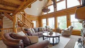 Ein Holzblockhaus-Interieur mit einer Holztreppe, Ledersofas, einem runden Couchtisch und großen Fenstern.