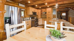 Ein Holzblockhaus-Interieur mit einer Küche, die einen Edelstahl-Kühlschrank, eine Mikrowelle und einen Backofen beinhaltet. Ein Esstisch mit Stühlen und einem Topf mit Pflanze darauf ist ebenfalls vorhanden.