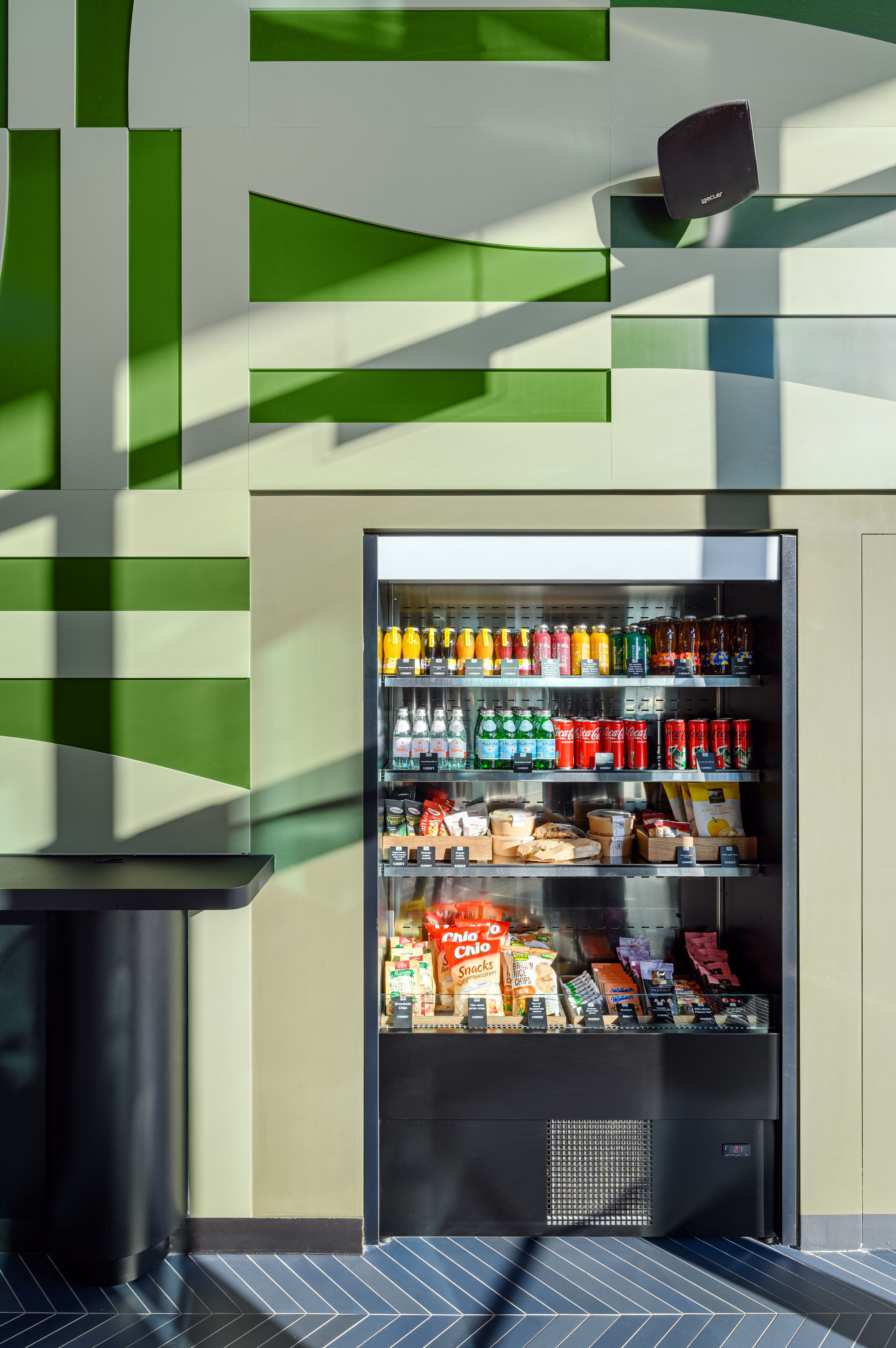 A vending machine stocked with beverages and snacks, located in a modern interior with geometric wall patterns and a tiled floor.