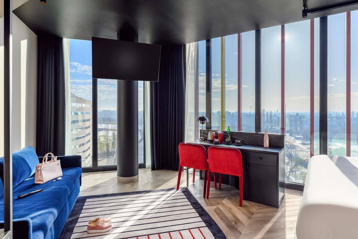 A modern hotel room with a large window, a blue sofa, a small desk with red chairs, and a TV mounted on the wall.