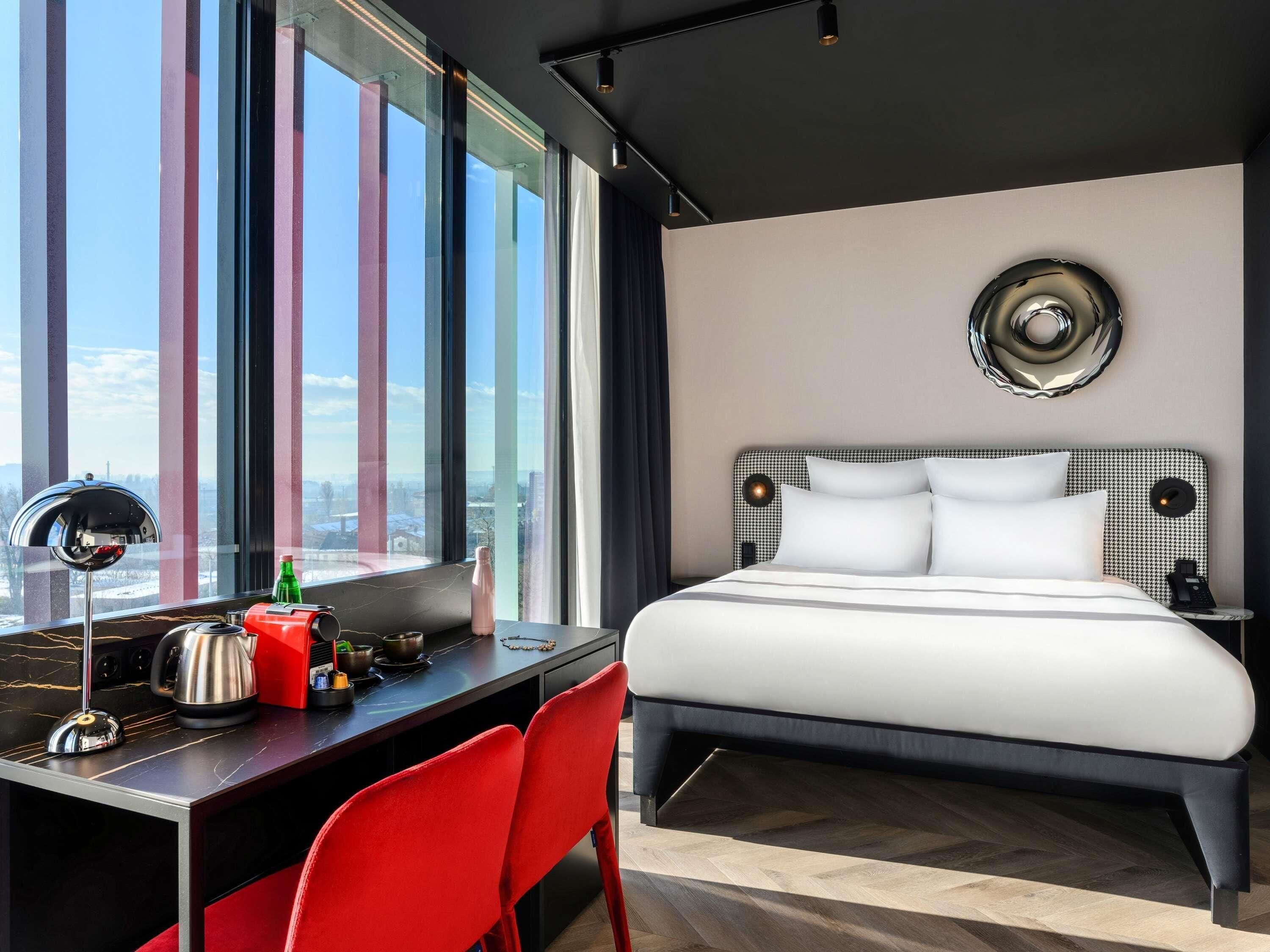 Una habitación de hotel moderna con una cama grande, un escritorio con una silla y vistas a la ciudad a través de amplias ventanas. Se ve un paisaje urbano al exterior.