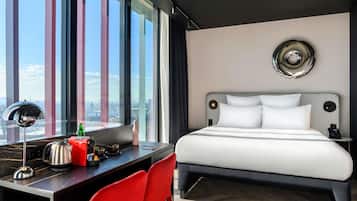 Una habitación de hotel moderna con una cama grande, un escritorio con una silla y vistas a la ciudad a través de amplias ventanas. Se ve un paisaje urbano al exterior.