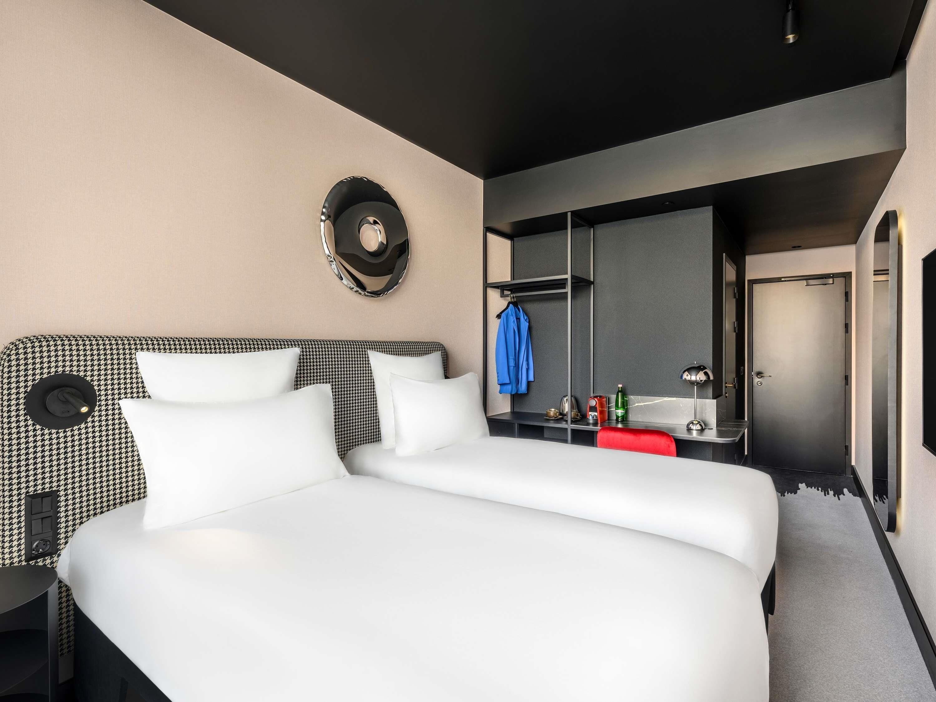 A modern hotel room with a large bed, a black and white headboard, a mirror, a small table with toiletries, and a door leading to a bathroom.