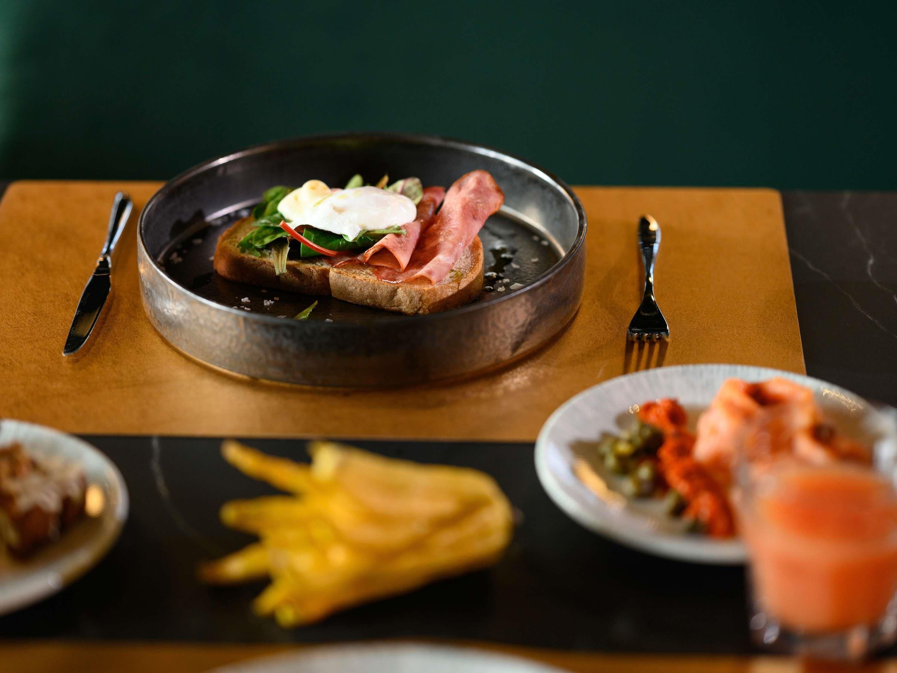 Une table dressée pour le petit-déjeuner avec des toasts, du jambon et un œuf poché sur une assiette, accompagnés de frites et d'une boisson.