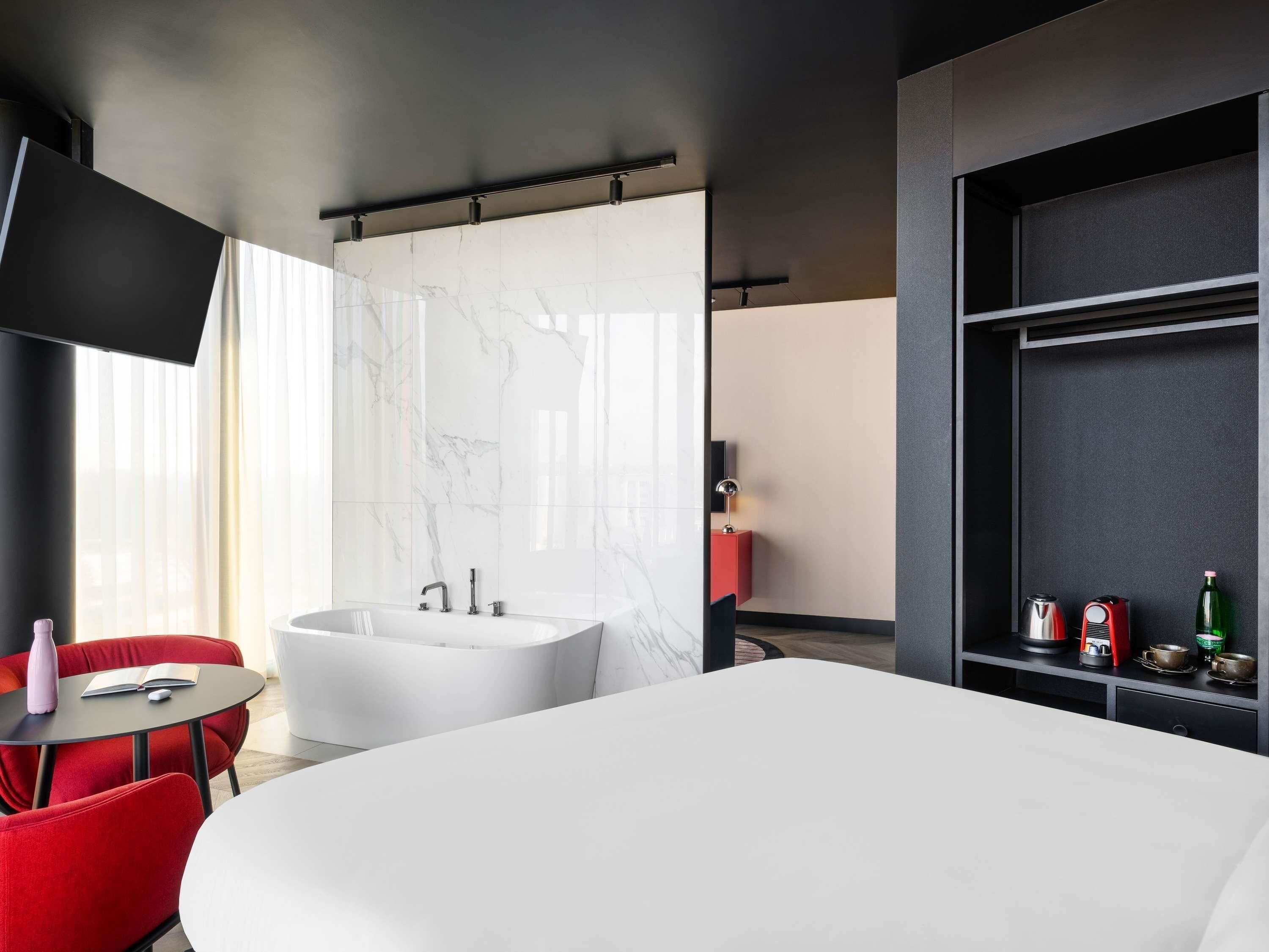 Habitación de hotel moderna con una cama grande, una zona de estar con una silla roja, una bañera blanca y un estante con una tetera y una botella.