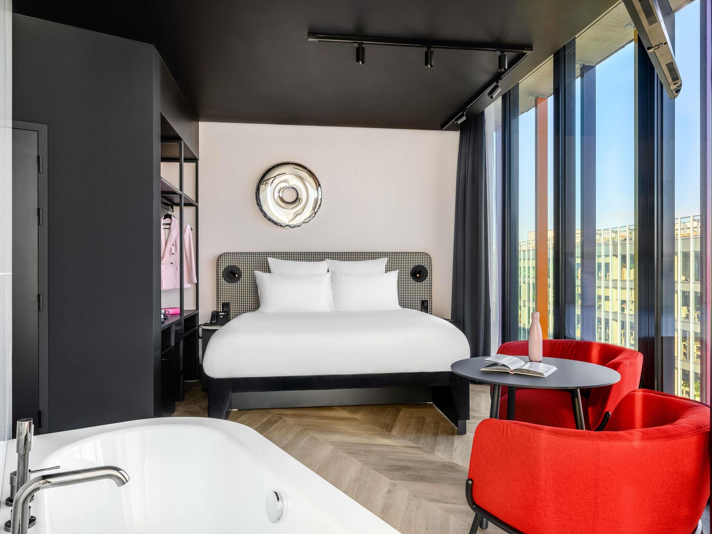 Een moderne hotelkamer met een groot bed, een zithoek met een rode fauteuil, een ronde tafel, een bad en uitzicht op de stad door grote ramen.