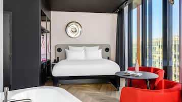 Camera d'albergo moderna con un letto grande, un'area salotto con una poltrona rossa, un tavolino rotondo, una vasca da bagno e vista sulla città attraverso ampie finestre.
