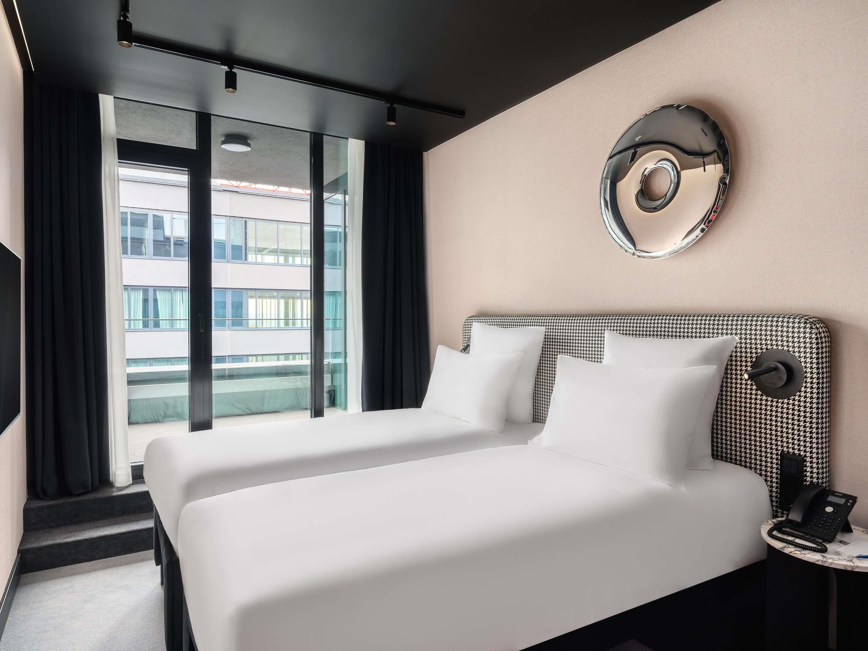A hotel room with a large bed, a round mirror, a telephone, and a view of a building through the window.
