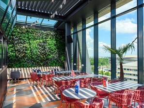 An outdoor dining area with tables and chairs, a green wall, and large windows. - Tribe Budapest Stadium (Budapest)