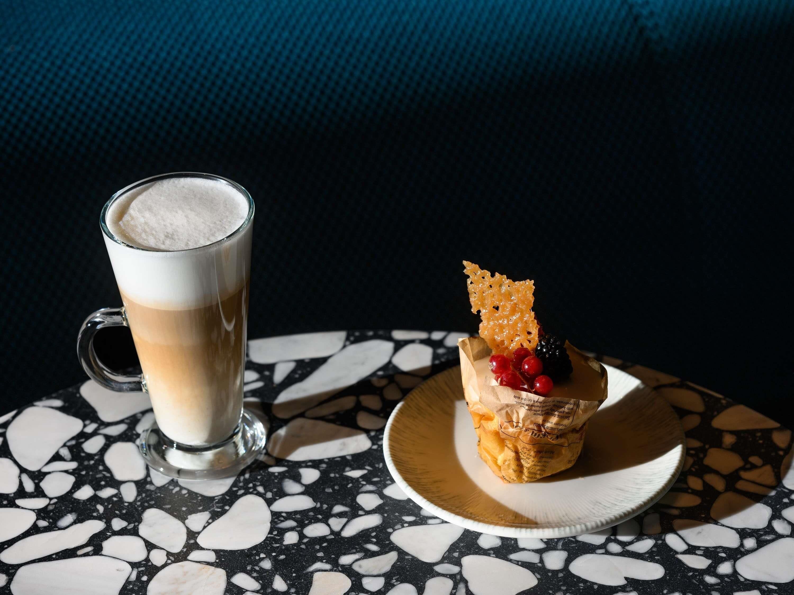 Una taza de café con espuma y un postre pequeño sobre un plato con forro de papel, sobre una mesa con vetas.