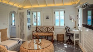 A cozy room with a round glass table, a wooden bench, and a wicker chair. There are two windows with views of the ocean, a door with a decorative lattice, and a small desk with a lamp. - Charming waterfront Caribbean estate - 2 min from pink sand beach (Little Harbour)