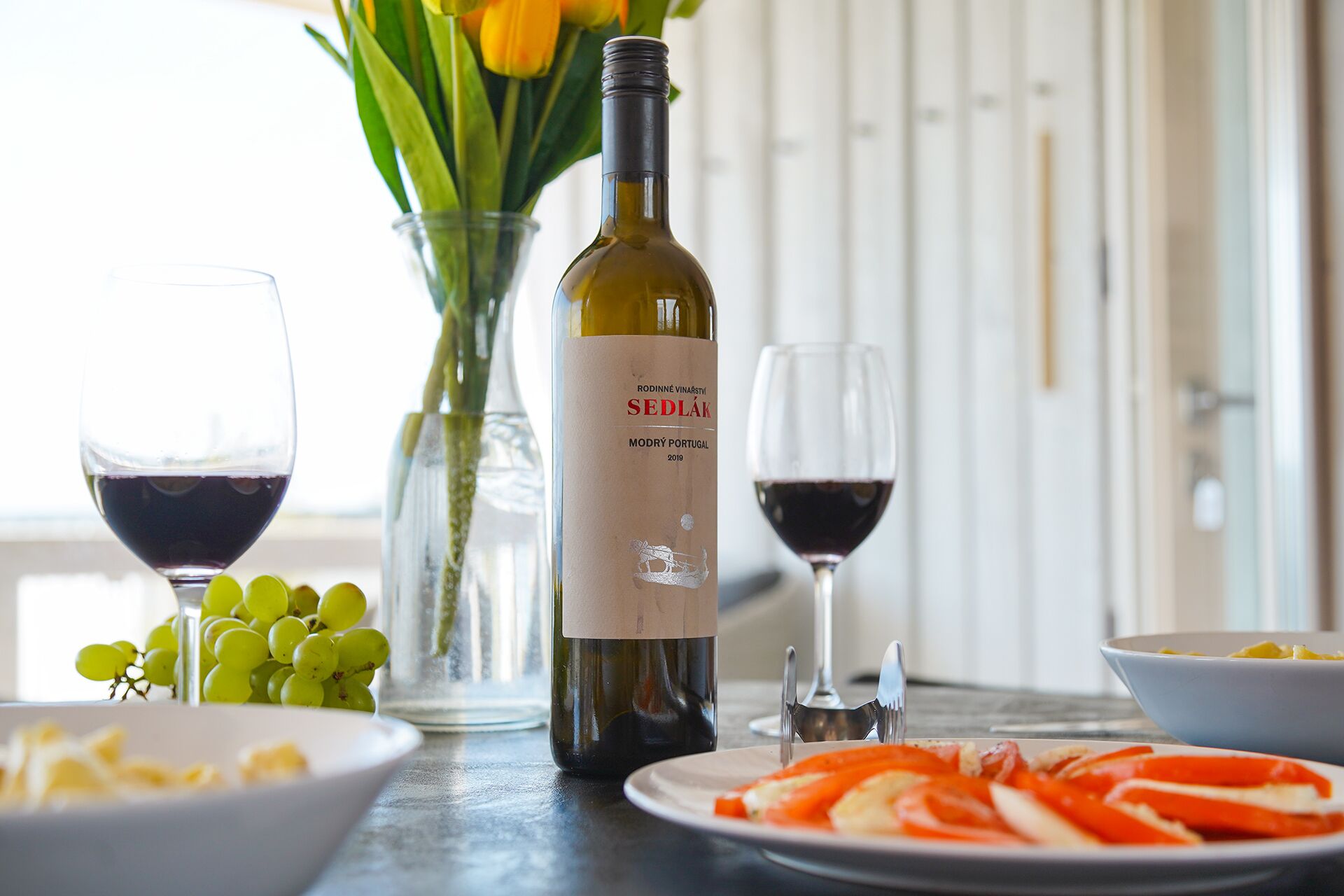 A table with a bottle of wine, glasses, a vase with flowers, and plates with food.