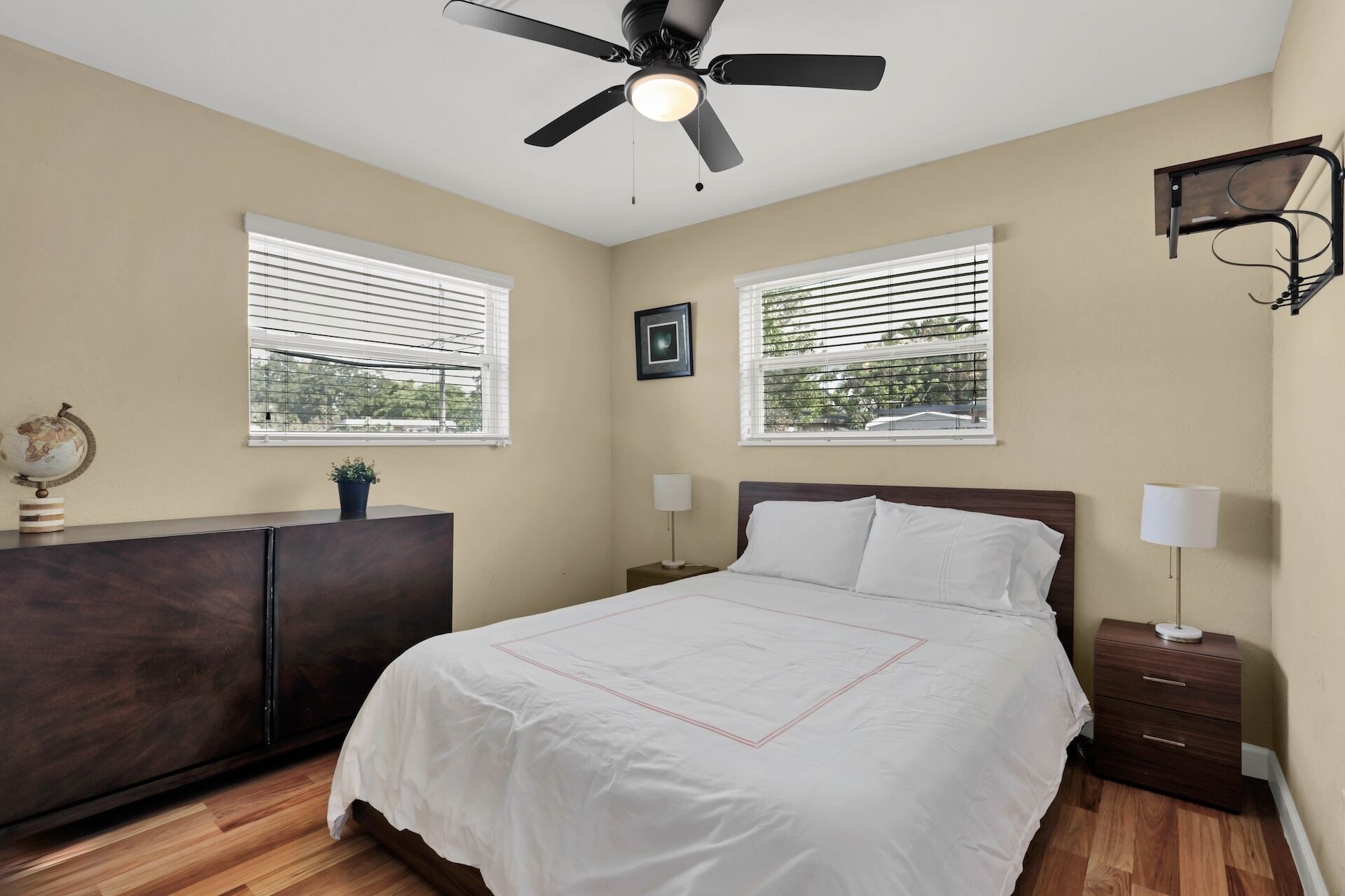 A bedroom with a bed, two bedside tables, a ceiling fan, and two windows with blinds.