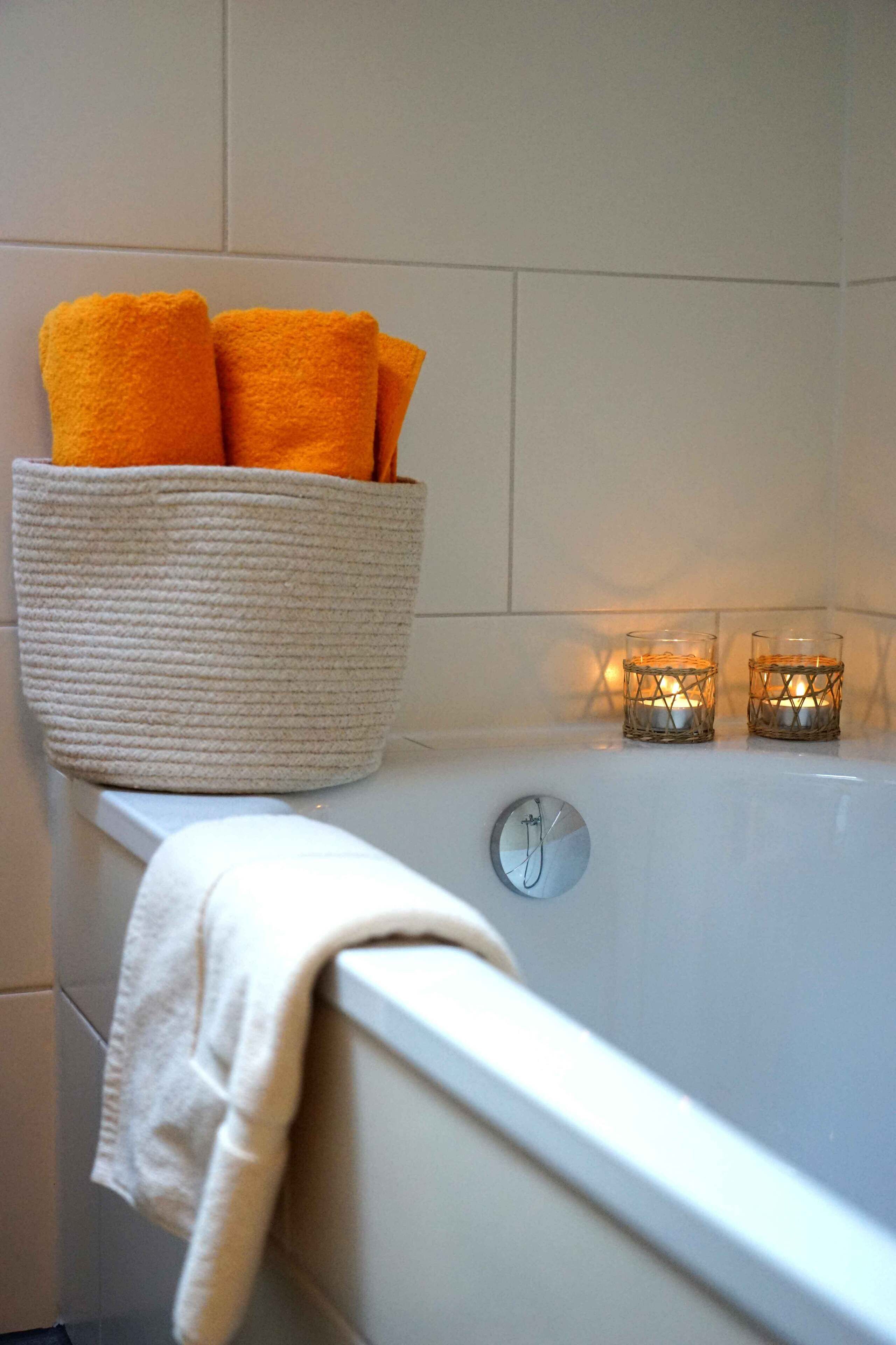A bathtub with a white towel and a basket of orange towels. There are two lit candles on the edge of the tub.