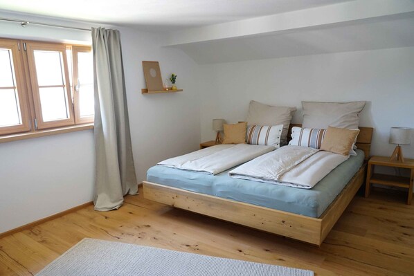 A bedroom with a wooden bed, a window with curtains, a bedside table with a lamp, and a small shelf with a picture and a plant.