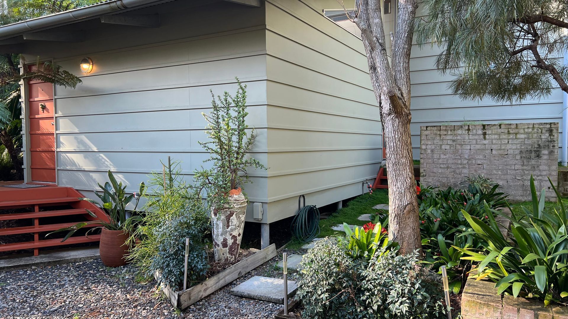 Une maison moderne avec un toit en pente, un petit porche et un jardin agrémenté de diverses plantes et d’un arbre.