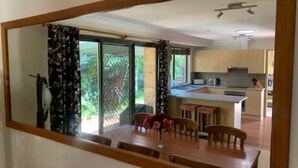 A dining area with a wooden table and chairs, a vase with flowers, and a view of a kitchen and outdoor greenery through a sliding door.