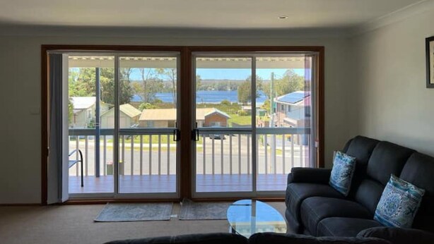 A living room with a black sofa, a glass coffee table, and a view through a sliding glass door to a deck with a railing. - Sanctuary 79 by Book Jervis Bay - Family home located in Sanctuary Point (Sanctuary Point)