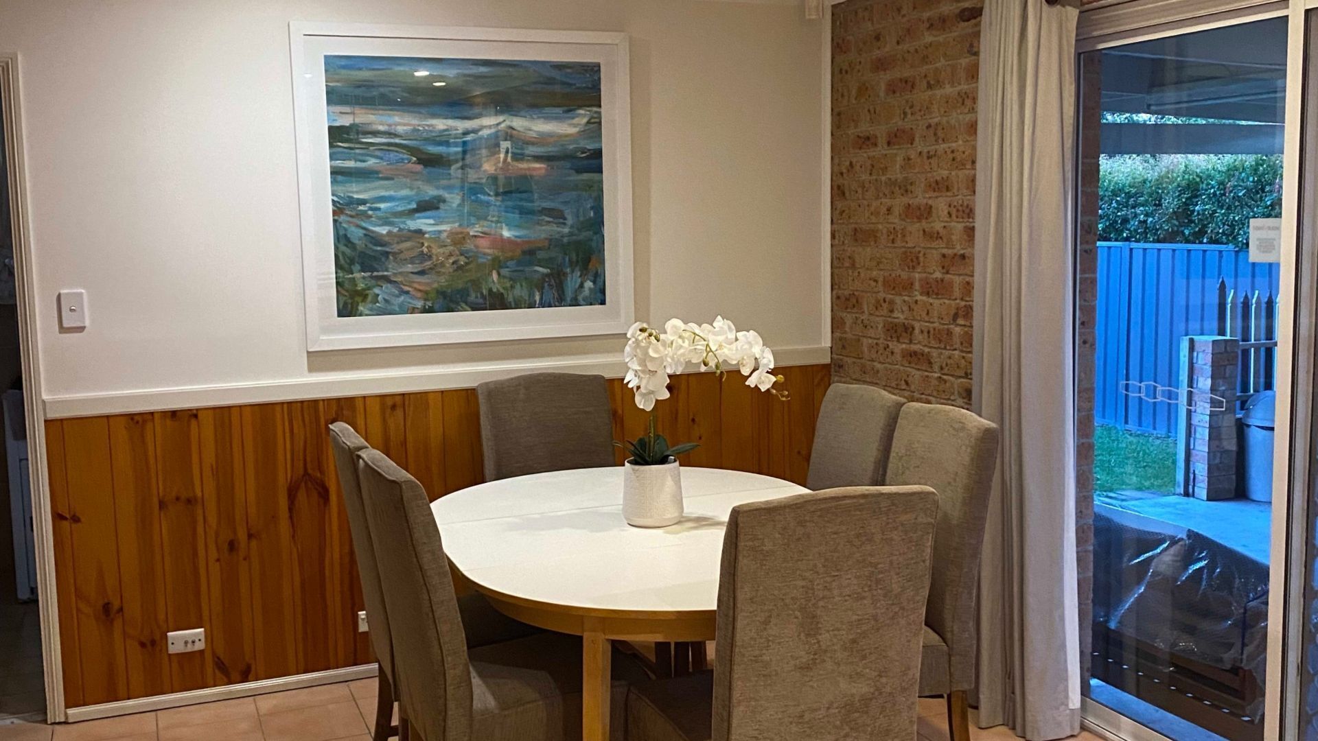 A dining area with a round table, chairs, a vase with white flowers, a painting on the wall, and a brick wall.
