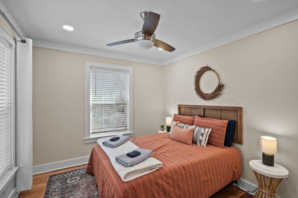 A bedroom with a bed, a ceiling fan, a window with blinds, a bedside lamp, and a small rug.