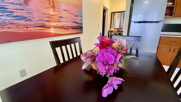 Un comedor con una mesa, sillas, un jarrón de flores, un cuadro de un atardecer y armarios de cocina.