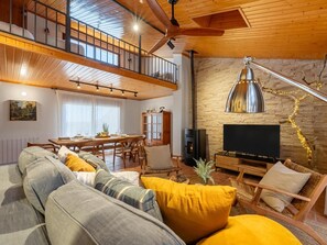A cozy living room with a high ceiling, wooden beams, a dining area, and a stone wall.