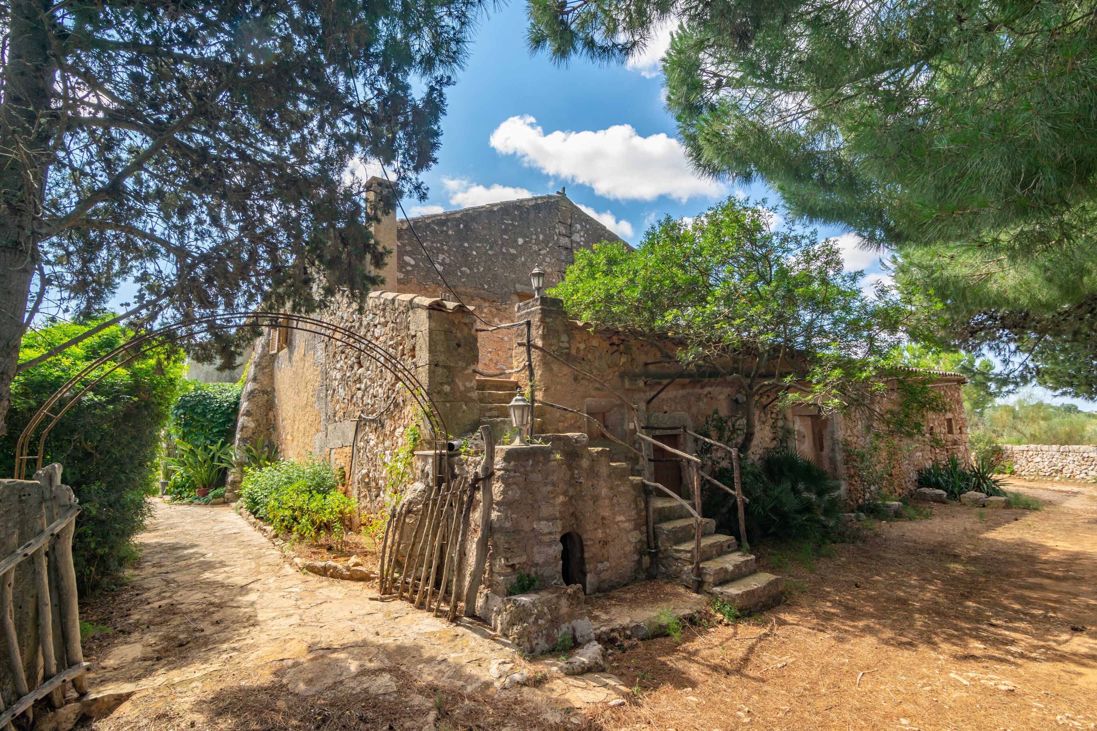 Un rústico edificio de piedra con una escalera que lleva a una puerta, rodeado de árboles y un sendero de tierra.