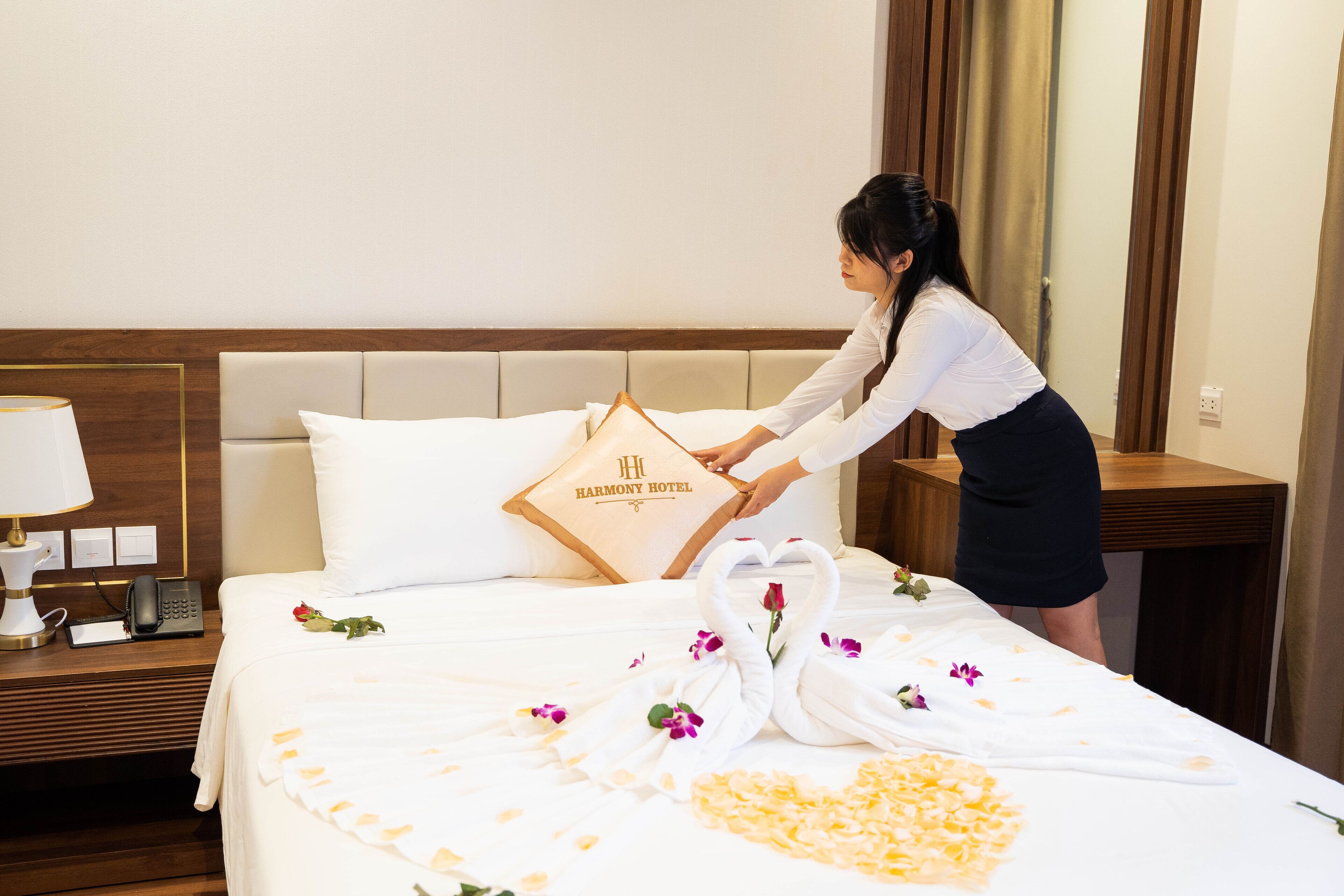 A hotel room with a neatly made bed, a pillow with the hotel name, a bedside phone, and a flower arrangement on the bed.