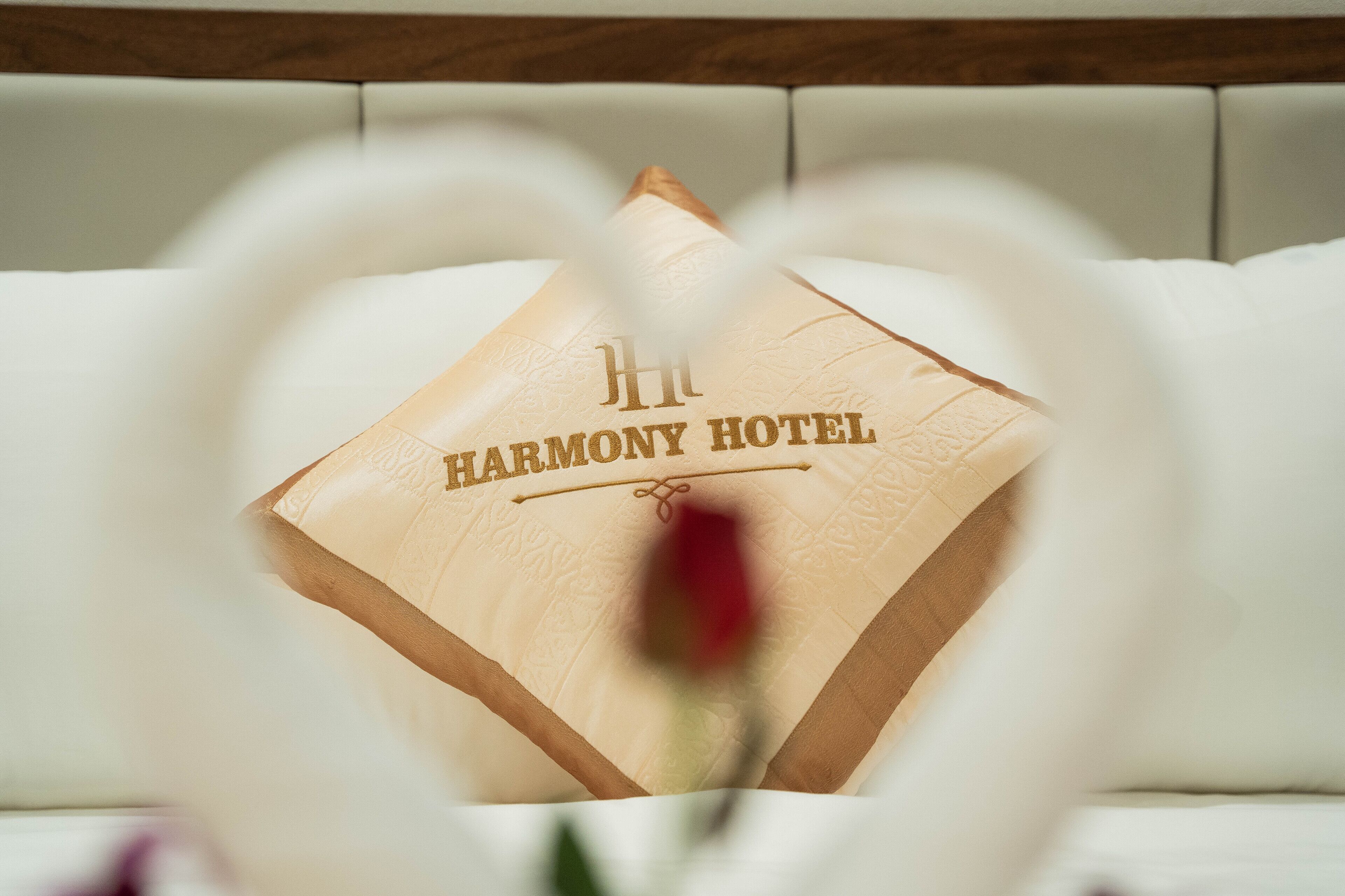 Una cama con un almohadón que dice "HARMONY HOTEL" y una flor roja desenfocada en primer plano.