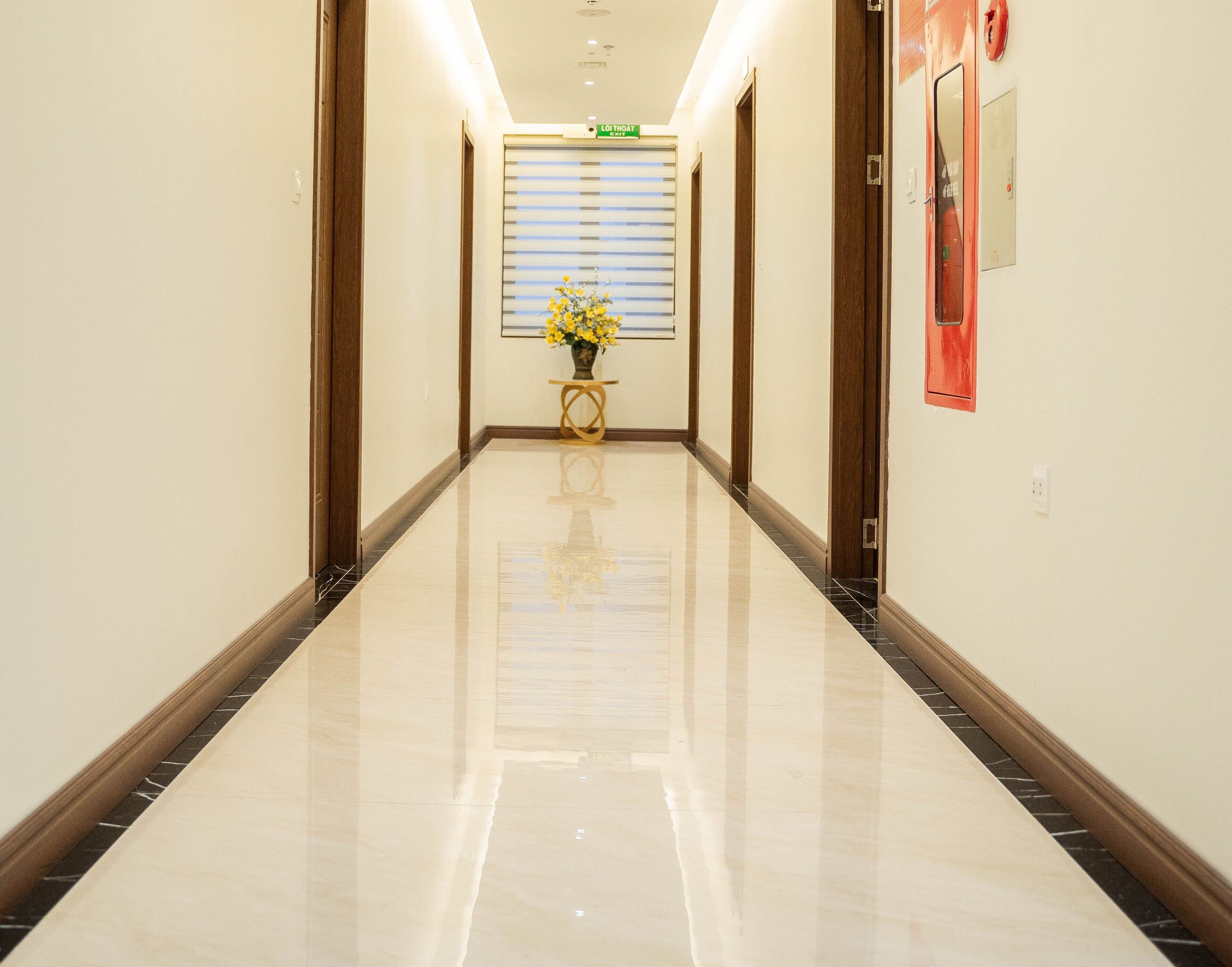 Un pasillo de hotel con piso pulido, puertas de madera y un florero con flores amarillas sobre una mesita.
