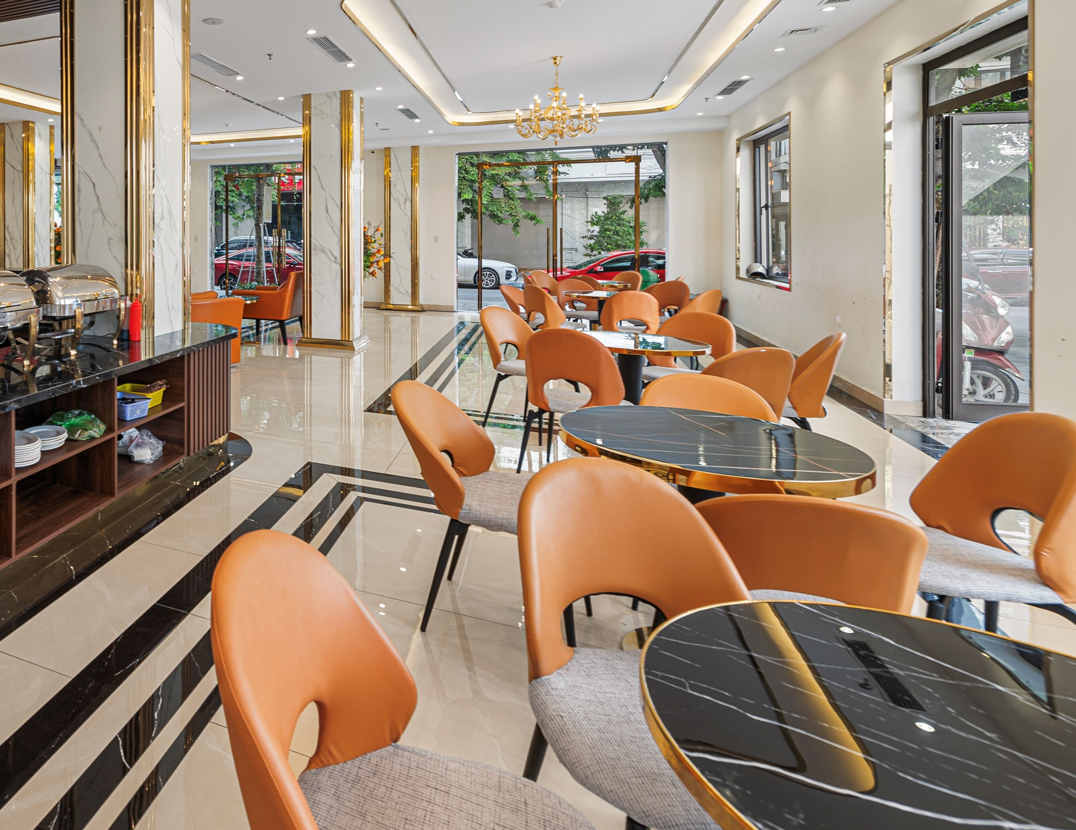 A modern hotel lobby with marble floors, orange chairs, and a central chandelier.