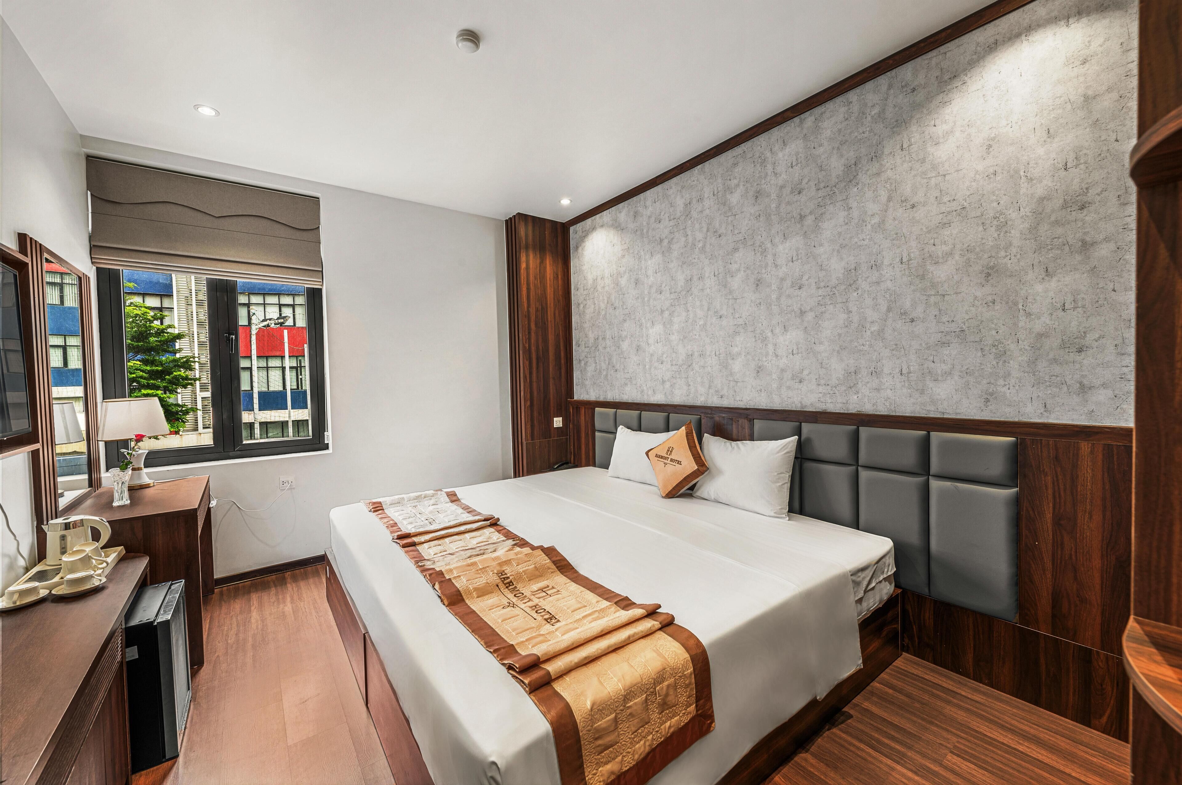 Una habitación de hotel moderna con una cama grande, un cabecero de madera, un escritorio con lámpara y un ventanal con vistas a edificios.