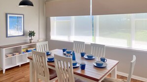 A dining area with a wooden table set for four, white chairs, and a large window with blinds.