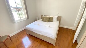 A bedroom with a wooden floor, a bed with white bedding, and a window with blinds.