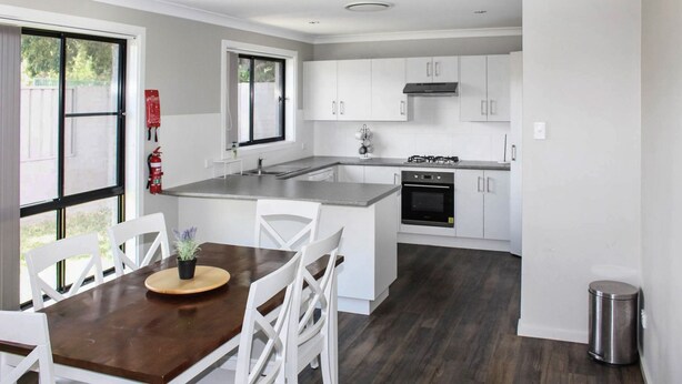 A modern kitchen with white cabinets, a central island, and a dining area with a wooden table and white chairs. - Quintet 1-157 - North Tamworth (Tamworth)