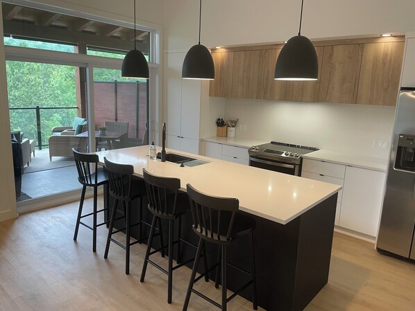 A modern kitchen with a large island, black bar stools, pendant lighting, and a view to an outdoor patio. - Beautiful Condo in Orford facing the mountain, pet-friendly (Orford)