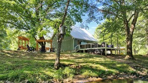 A rustic cabin with a porch and outdoor seating, surrounded by lush greenery and a small stream. - Creekfront! 5* Views, hot tub, fire pit, huge deck (Blue Ridge)