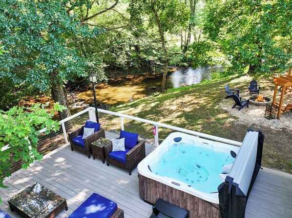 Una terraza con mobiliario exterior, un jacuzzi y un área con fogata, todo rodeado de árboles.